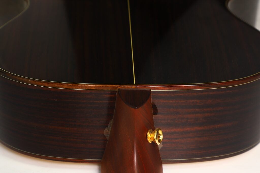 Nahaufnahme der Rückseite und der Seite einer Taylor 712 Akustikgitarre, die die dunkle Holzmaserung, die gelbe Einfassung und den mit einem goldenen Gurtknopf versehenen Halsfuß hervorhebt. Nahaufnahme der Rückseite und der Seite einer Taylor 712 Akustikgitarre, die die dunkle Holzmaserung, die gelbe Einfassung und den mit einem goldenen Gurtknopf versehenen Halsfuß hervorhebt.