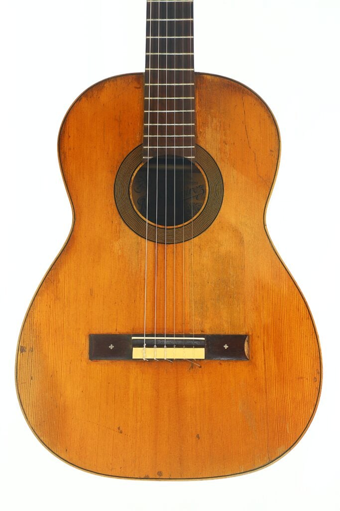 Eine Nahaufnahme einer José-Ramirez-Klassikgitarre aus dem Jahr 1932 mit einem abgenutzten Holzkorpus, sechs Saiten und sichtbaren Kratzern, die Spuren häufiger Nutzung aufweisen. Eine Nahaufnahme einer José-Ramirez-Klassikgitarre aus dem Jahr 1932 mit einem abgenutzten Holzkorpus, sechs Saiten und sichtbaren Kratzern, die Spuren häufiger Nutzung aufweisen.