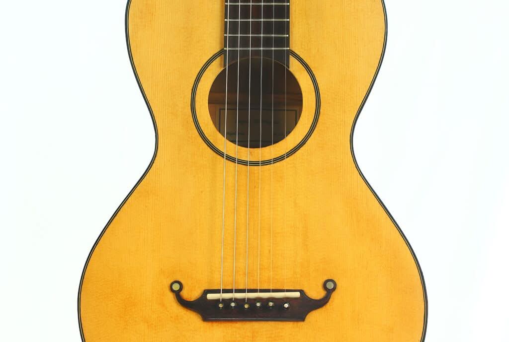 Eine Nahaufnahme der Lacote Stil Romantikgitarre zeigt ihren hellen Holzkorpus, die sechs Saiten, das runde Schallloch und den dunklen Steg, alles vor einem schlichten weißen Hintergrund. Eine Nahaufnahme der Lacote Stil Romantikgitarre zeigt ihren hellen Holzkorpus, die sechs Saiten, das runde Schallloch und den dunklen Steg, alles vor einem schlichten weißen Hintergrund.