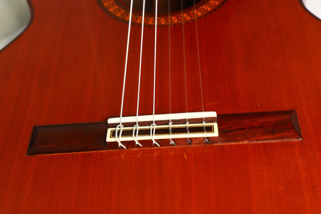 Nahaufnahme des Stegs einer José Ramirez 1a 1973 Klassische Gitarre, auf der sechs Saiten zu sehen sind, die am polierten, rotbraunen Holzkorpus mit einem weißen Sattel befestigt sind. Nahaufnahme des Stegs einer José Ramirez 1a 1973 Klassische Gitarre, auf der sechs Saiten zu sehen sind, die am polierten, rotbraunen Holzkorpus mit einem weißen Sattel befestigt sind.