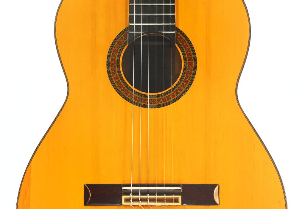 Nahaufnahme des Korpus einer Flamenco-Gitarre von Sobrinos de Santos Hernandez aus dem Jahr 1974 mit sechs Saiten, einer dekorativen Rosette um das Schallloch und einem Steg aus Holz vor weißem Hintergrund. Nahaufnahme des Korpus einer Flamenco-Gitarre von Sobrinos de Santos Hernandez aus dem Jahr 1974 mit sechs Saiten, einer dekorativen Rosette um das Schallloch und einem Steg aus Holz vor weißem Hintergrund.