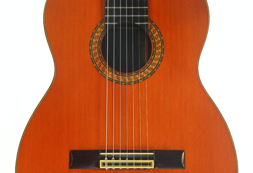 Eine Nahaufnahme des Korpus einer 7-saitigen klassischen Gitarre vom Typ „Ramirez Stil“ von Melchor Rodriguez aus dem Jahr 1980, die sieben Saiten, einen Steg aus Holz, eine verzierte Rosette um das Schallloch sowie eine polierte braune Lackierung zeigt. Eine Nahaufnahme des Korpus einer 7-saitigen klassischen Gitarre vom Typ „Ramirez Stil“ von Melchor Rodriguez aus dem Jahr 1980, die sieben Saiten, einen Steg aus Holz, eine verzierte Rosette um das Schallloch sowie eine polierte braune Lackierung zeigt.