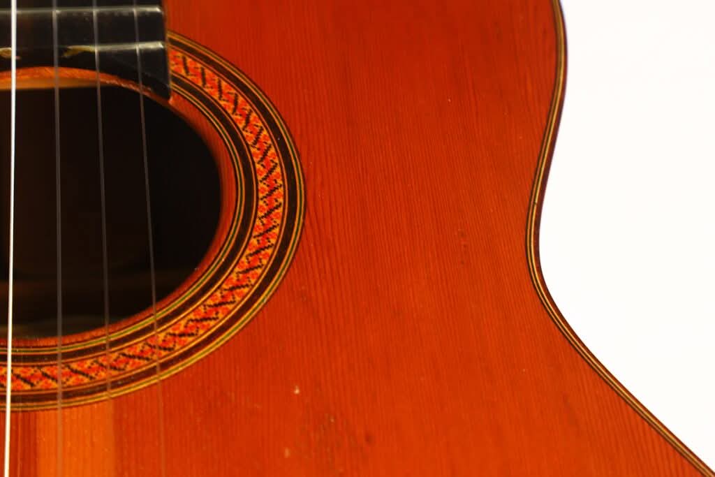 Nahaufnahme der „Antonio Duran 1974 Klassische Gitarre“, die die feine Holzmaserung des Korpus der klassischen Akustikgitarre, einen Teil des Schalllochs mit seiner dekorativen Rosette sowie die Gitarrensaiten vor einem weißen Hintergrund hervorhebt. Nahaufnahme der „Antonio Duran 1974 Klassische Gitarre“, die die feine Holzmaserung des Korpus der klassischen Akustikgitarre, einen Teil des Schalllochs mit seiner dekorativen Rosette sowie die Gitarrensaiten vor einem weißen Hintergrund hervorhebt.