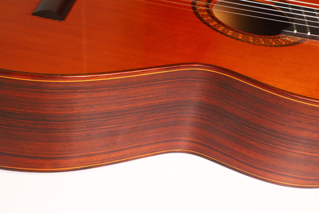 Nahaufnahme der Seite und eines Teils der Oberseite einer José Ramirez 1a 1973 Klassische Gitarre, die die wunderschöne Holzmaserung, die sanften Rundungen und die Saiten zeigt. Das Bild verdeutlicht die außergewöhnliche Handwerkskunst und die reichhaltige Textur dieses klassischen Instruments. Nahaufnahme der Seite und eines Teils der Oberseite einer José Ramirez 1a 1973 Klassische Gitarre, die die wunderschöne Holzmaserung, die sanften Rundungen und die Saiten zeigt. Das Bild verdeutlicht die außergewöhnliche Handwerkskunst und die reichhaltige Textur dieses klassischen Instruments.