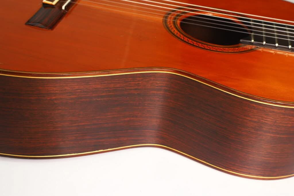 Nahaufnahme der Seite und Oberseite der „Klassische Gitarre“ von Antonio Duran aus dem Jahr 1974, die die Holzmaserung, die dekorative Rosette um das Schallloch sowie einen Teil der Saiten und des Stegs vor weißem Hintergrund zeigt. Nahaufnahme der Seite und Oberseite der „Klassische Gitarre“ von Antonio Duran aus dem Jahr 1974, die die Holzmaserung, die dekorative Rosette um das Schallloch sowie einen Teil der Saiten und des Stegs vor weißem Hintergrund zeigt.