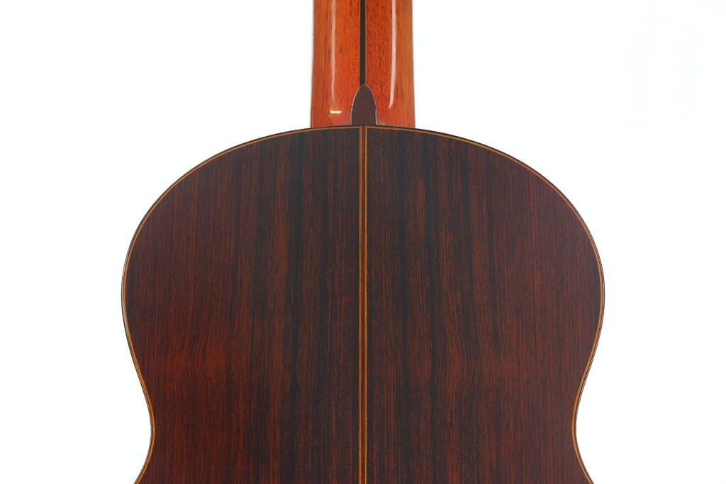 Nahaufnahme der Rückseite einer 7-saitigen klassischen Gitarre vom Typ „Ramirez Stil“ von Melchor Rodriguez aus dem Jahr 1980, die die dunkle Holzmaserung und eine vertikale Mittelnaht hervorhebt; oben ist vor weißem Hintergrund ein Teil des Halses zu sehen. Nahaufnahme der Rückseite einer 7-saitigen klassischen Gitarre vom Typ „Ramirez Stil“ von Melchor Rodriguez aus dem Jahr 1980, die die dunkle Holzmaserung und eine vertikale Mittelnaht hervorhebt; oben ist vor weißem Hintergrund ein Teil des Halses zu sehen.