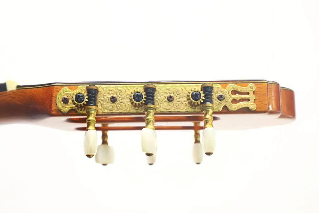 Nahaufnahme der Stimmwirbel und der verzierten Messing-Stimmplatte am Kopf der „Gran Klassische Gitarre“ von Raimundo y Aparicio Corujo vor einem schlichten weißen Hintergrund. Nahaufnahme der Stimmwirbel und der verzierten Messing-Stimmplatte am Kopf der „Gran Klassische Gitarre“ von Raimundo y Aparicio Corujo vor einem schlichten weißen Hintergrund.