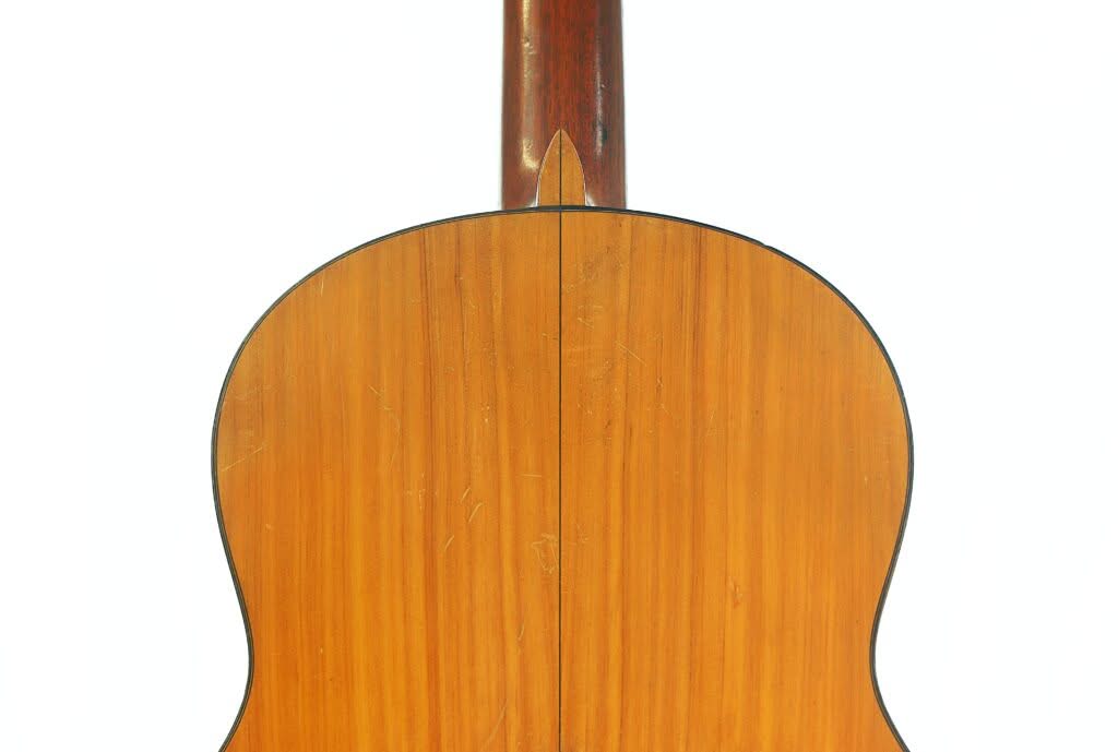 Nahaufnahme der Rückseite einer Flamenco-Gitarre von Manuel de la Chica aus dem Jahr 1954, die eine glatte, polierte Holzoberfläche mit einer Naht in der Mitte und leichten Kratzern aufweist; der Hals verläuft nach oben. Nahaufnahme der Rückseite einer Flamenco-Gitarre von Manuel de la Chica aus dem Jahr 1954, die eine glatte, polierte Holzoberfläche mit einer Naht in der Mitte und leichten Kratzern aufweist; der Hals verläuft nach oben.
