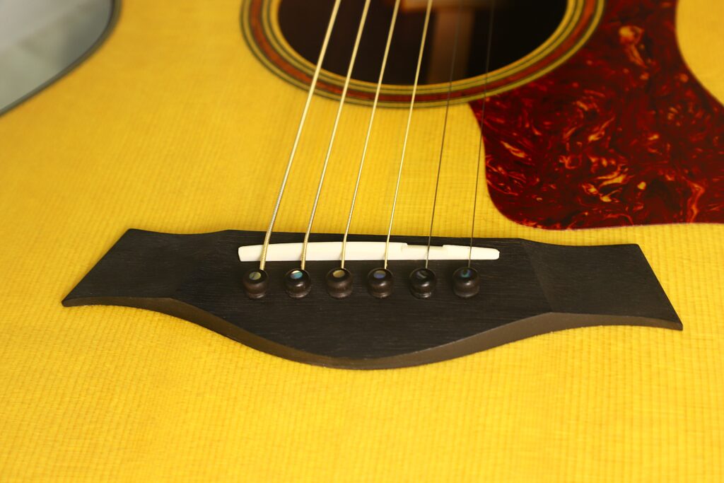 Nahaufnahme des Stegs der Taylor 712 Akustikgitarre, auf der die Saiten, die Stegstifte und ein Teil des gelben Holzkörpers der Gitarre mit einem braunen Schlagbrett zu sehen sind. Nahaufnahme des Stegs der Taylor 712 Akustikgitarre, auf der die Saiten, die Stegstifte und ein Teil des gelben Holzkörpers der Gitarre mit einem braunen Schlagbrett zu sehen sind.