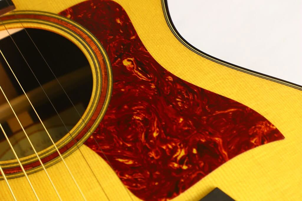 Nahaufnahme des Korpus der Taylor 712 Akustikgitarre, die die gelbe Holzmaserung, das runde Schallloch und ein rotes Schlagbrett mit Schildpatt-Muster zeigt. Nahaufnahme des Korpus der Taylor 712 Akustikgitarre, die die gelbe Holzmaserung, das runde Schallloch und ein rotes Schlagbrett mit Schildpatt-Muster zeigt.