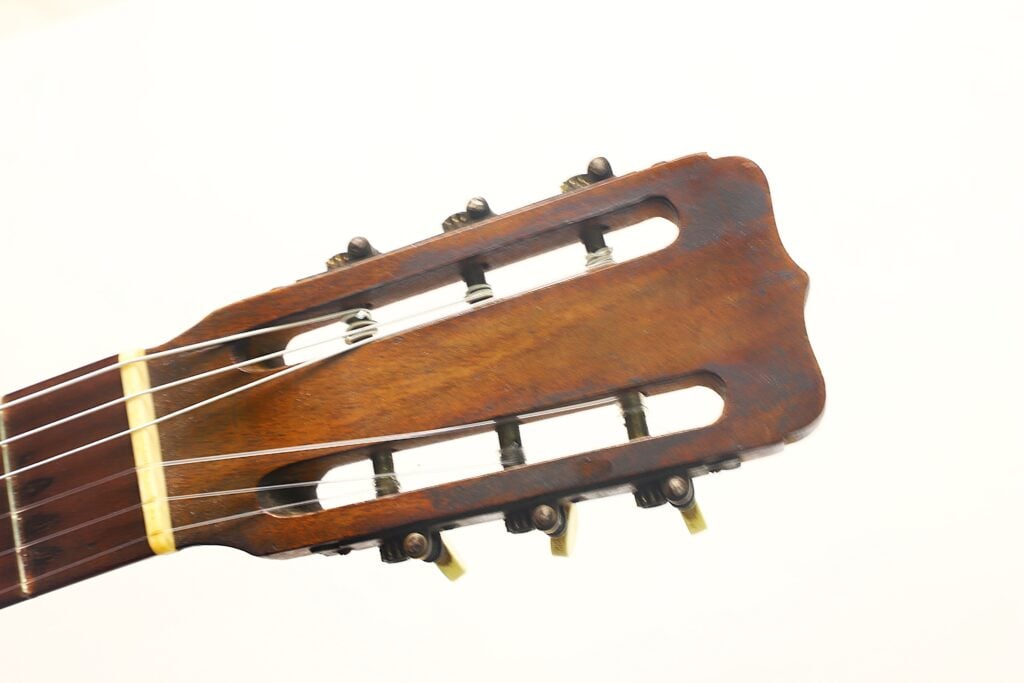 Nahaufnahme des Kopfes einer „José Ramirez 1932 Klassische Gitarre“ mit sechs Stimmwirbeln und Nylonsaiten vor einem schlichten weißen Hintergrund. Nahaufnahme des Kopfes einer „José Ramirez 1932 Klassische Gitarre“ mit sechs Stimmwirbeln und Nylonsaiten vor einem schlichten weißen Hintergrund.