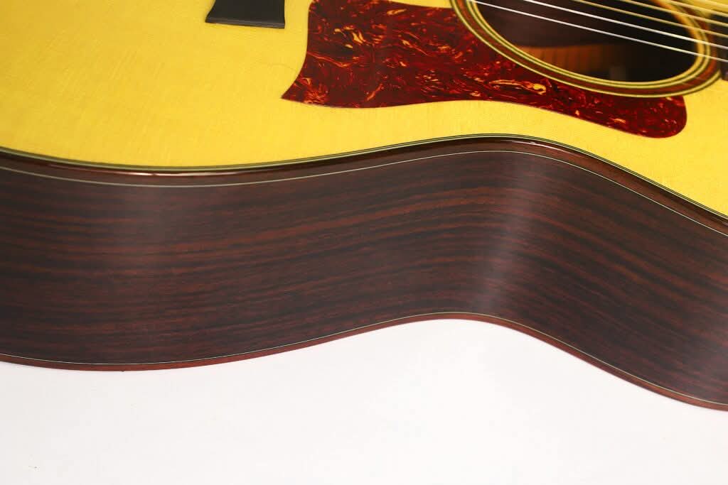 Nahaufnahme der Seite und des Korpus einer Taylor 712 Akustikgitarre mit Holzoberfläche und sichtbarer Maserung sowie Teilen der Saiten und des Schlagbretts vor weißem Hintergrund. Nahaufnahme der Seite und des Korpus einer Taylor 712 Akustikgitarre mit Holzoberfläche und sichtbarer Maserung sowie Teilen der Saiten und des Schlagbretts vor weißem Hintergrund.