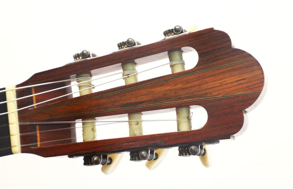 Nahaufnahme des Kopfes der „Hauser Klassische Gitarre“ von Alastair McNeil aus dem Jahr 1997 mit Holzoberfläche, jeweils drei weißen Stimmwirbeln auf jeder Seite und Nylonsaiten vor weißem Hintergrund. Nahaufnahme des Kopfes der „Hauser Klassische Gitarre“ von Alastair McNeil aus dem Jahr 1997 mit Holzoberfläche, jeweils drei weißen Stimmwirbeln auf jeder Seite und Nylonsaiten vor weißem Hintergrund.