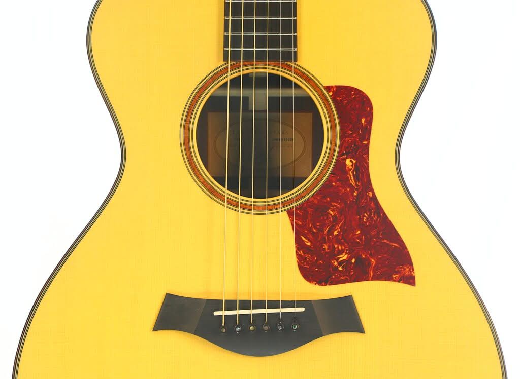 Nahaufnahme des Korpus und der Saiten einer Taylor 712 Akustikgitarre, mit Blick auf das Schallloch, den Steg und ein rot marmoriertes Schlagbrett, das sich vom hellen Holzfarbton abhebt. Nahaufnahme des Korpus und der Saiten einer Taylor 712 Akustikgitarre, mit Blick auf das Schallloch, den Steg und ein rot marmoriertes Schlagbrett, das sich vom hellen Holzfarbton abhebt.