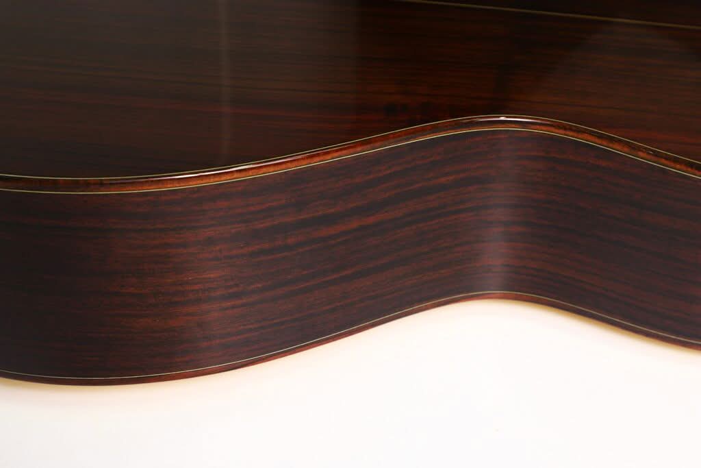Nahaufnahme der Seite der Taylor 712 Akustikgitarre, die die glatte, dunkle Holzmaserung und die sanft geschwungene Form vor einem hellen Hintergrund hervorhebt. Nahaufnahme der Seite der Taylor 712 Akustikgitarre, die die glatte, dunkle Holzmaserung und die sanft geschwungene Form vor einem hellen Hintergrund hervorhebt.
