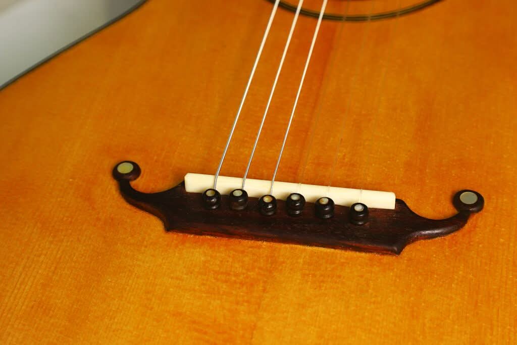 Nahaufnahme des Stegs und der Saiten einer akustischen „Lacote Stil Romantikgitarre“ auf einem hellen Holzkorpus, wobei sechs Saitenstifte, die die Saiten fixieren, sowie ein Teil der Decke zu sehen sind. Nahaufnahme des Stegs und der Saiten einer akustischen „Lacote Stil Romantikgitarre“ auf einem hellen Holzkorpus, wobei sechs Saitenstifte, die die Saiten fixieren, sowie ein Teil der Decke zu sehen sind.