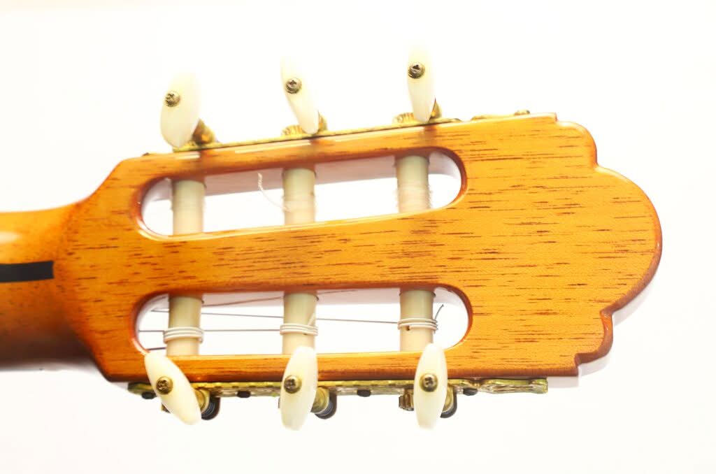 Nahaufnahme des Kopfes der „Gran Klassische Gitarre“ von Raimundo y Aparicio Corujo mit sechs weißen Stimmwirbeln, Saiten und hellbrauner Holzmaserung vor weißem Hintergrund. Nahaufnahme des Kopfes der „Gran Klassische Gitarre“ von Raimundo y Aparicio Corujo mit sechs weißen Stimmwirbeln, Saiten und hellbrauner Holzmaserung vor weißem Hintergrund.