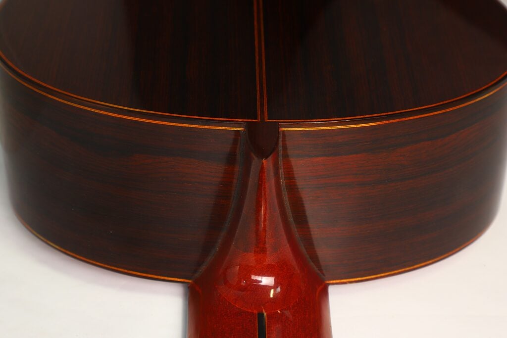 Nahaufnahme des Halsansatzes der 7-saitigen klassischen Gitarre „Ramirez Stil“ von Melchor Rodriguez aus dem Jahr 1980, die auf weißem Hintergrund poliertes dunkles Holz mit helleren Zierleisten und einer glatten, glänzenden Oberfläche zeigt. Nahaufnahme des Halsansatzes der 7-saitigen klassischen Gitarre „Ramirez Stil“ von Melchor Rodriguez aus dem Jahr 1980, die auf weißem Hintergrund poliertes dunkles Holz mit helleren Zierleisten und einer glatten, glänzenden Oberfläche zeigt.