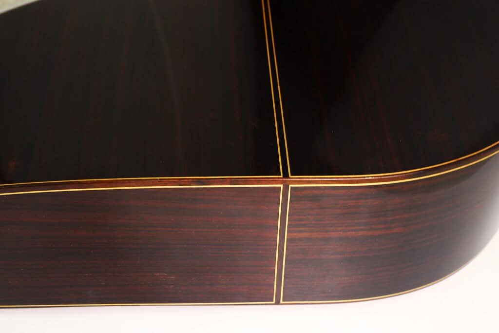 Nahaufnahme einer Ecke der „Gran Klassische Gitarre“ von Raimundo y Aparicio Corujo, die den dunklen Holzkorpus mit gelber Zierleiste und sichtbarer Holzmaserung hervorhebt und die detailreiche Handwerkskunst sowie die glatte, glänzende Oberfläche zur Geltung bringt. Nahaufnahme einer Ecke der „Gran Klassische Gitarre“ von Raimundo y Aparicio Corujo, die den dunklen Holzkorpus mit gelber Zierleiste und sichtbarer Holzmaserung hervorhebt und die detailreiche Handwerkskunst sowie die glatte, glänzende Oberfläche zur Geltung bringt.
