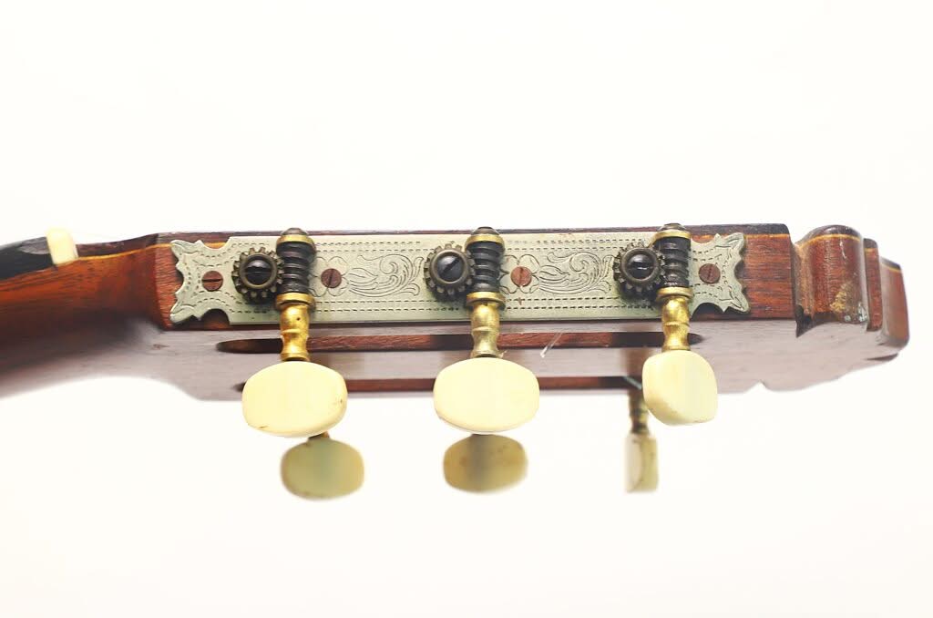 Nahaufnahme der Seite des Kopfes einer Flamenco-Gitarre von Manuel de la Chica aus dem Jahr 1954, die drei Stimmwirbel mit cremefarbenen Knöpfen und kunstvoll verzierten Metallteilen am Mechanismus vor weißem Hintergrund zeigt. Nahaufnahme der Seite des Kopfes einer Flamenco-Gitarre von Manuel de la Chica aus dem Jahr 1954, die drei Stimmwirbel mit cremefarbenen Knöpfen und kunstvoll verzierten Metallteilen am Mechanismus vor weißem Hintergrund zeigt.