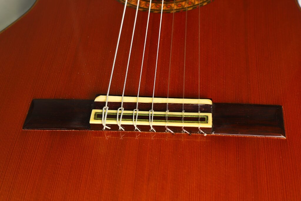 Nahaufnahme des Stegs und der Saiten einer 7-saitigen klassischen Gitarre vom Typ „Ramirez Stil“ von Melchor Rodriguez aus dem Jahr 1980 mit braunem Holzkorpus, auf der sieben am Steg befestigte Nylonsaiten zu sehen sind. Nahaufnahme des Stegs und der Saiten einer 7-saitigen klassischen Gitarre vom Typ „Ramirez Stil“ von Melchor Rodriguez aus dem Jahr 1980 mit braunem Holzkorpus, auf der sieben am Steg befestigte Nylonsaiten zu sehen sind.
