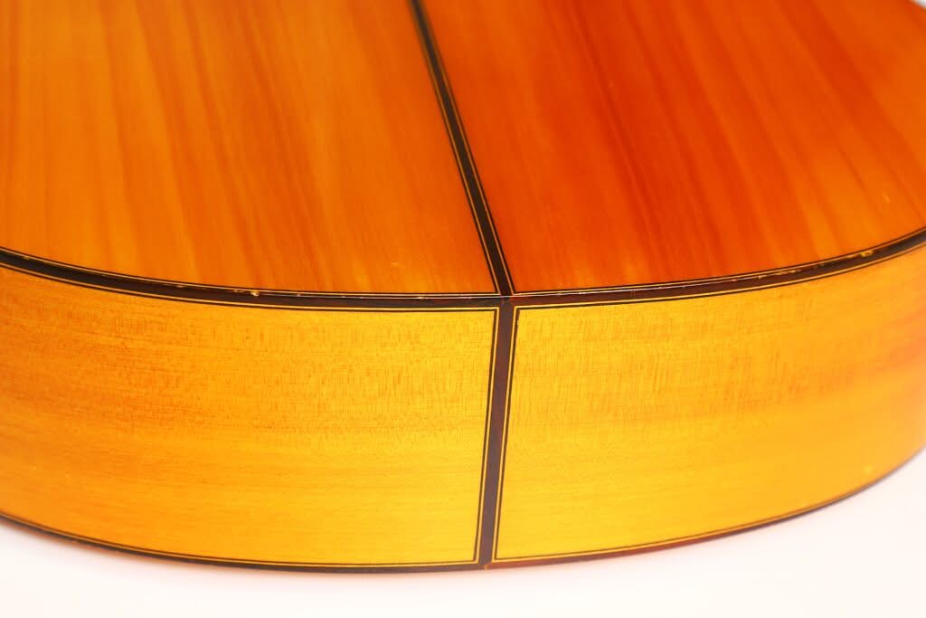 Nahaufnahme der Rückseite und der Seite einer Flamenco-Gitarre von Sobrinos de Santos Hernandez aus dem Jahr 1974, die vor einem sanften weißen Hintergrund die warme, polierte Holzmaserung, die dunkle Einfassung und die feine Handwerkskunst hervorhebt. Nahaufnahme der Rückseite und der Seite einer Flamenco-Gitarre von Sobrinos de Santos Hernandez aus dem Jahr 1974, die vor einem sanften weißen Hintergrund die warme, polierte Holzmaserung, die dunkle Einfassung und die feine Handwerkskunst hervorhebt.