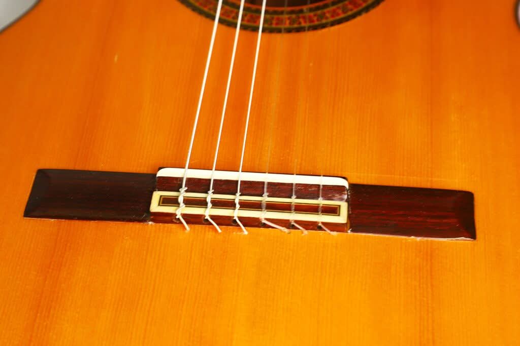 Nahaufnahme der Brücke und der Saiten einer Flamenco-Gitarre von Sobrinos de Santos Hernandez aus dem Jahr 1974, die den detailreichen Holzkorpus, die Saiten und den Sattel zeigt. Nahaufnahme der Brücke und der Saiten einer Flamenco-Gitarre von Sobrinos de Santos Hernandez aus dem Jahr 1974, die den detailreichen Holzkorpus, die Saiten und den Sattel zeigt.