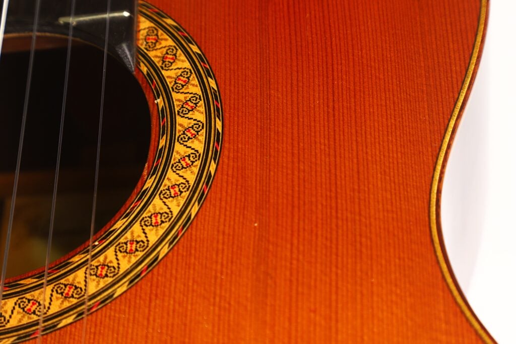 Nahaufnahme des Korpus der 7-saitigen klassischen Gitarre „Ramirez Stil“ von Melchor Rodriguez aus dem Jahr 1980, bei der die Saiten, das Schallloch und die filigranen Ziermuster rund um die Rosette besonders hervorgehoben sind. Nahaufnahme des Korpus der 7-saitigen klassischen Gitarre „Ramirez Stil“ von Melchor Rodriguez aus dem Jahr 1980, bei der die Saiten, das Schallloch und die filigranen Ziermuster rund um die Rosette besonders hervorgehoben sind.