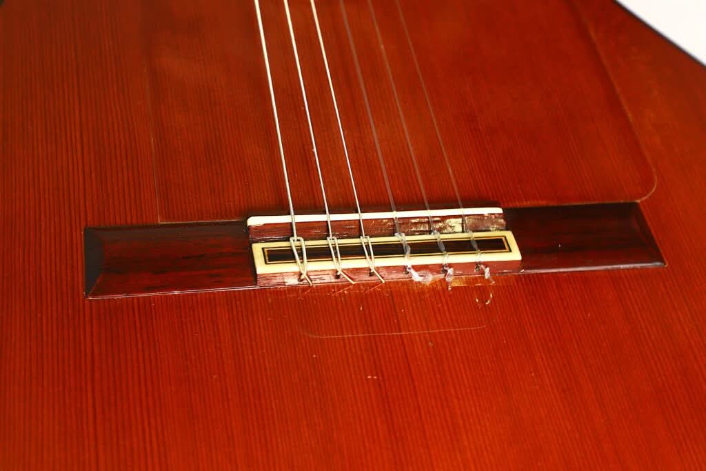 Nahaufnahme des Stegs und der Nylonsaiten auf dem polierten Holzkorpus der Jose Ramirez 1a 1968 Flamenco-Gitarre. Der Fokus liegt auf den Saiten und dem Stegbereich, wodurch die feinen Details und die handwerkliche Qualität dieses renommierten Instruments besonders hervorgehoben werden. Nahaufnahme des Stegs und der Nylonsaiten auf dem polierten Holzkorpus der Jose Ramirez 1a 1968 Flamenco-Gitarre. Der Fokus liegt auf den Saiten und dem Stegbereich, wodurch die feinen Details und die handwerkliche Qualität dieses renommierten Instruments besonders hervorgehoben werden.