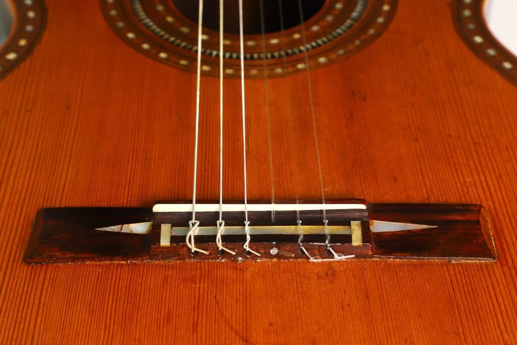 Nahaufnahme des Stegs und der Saiten einer „Salvador Ibanez 1900 Klassische Gitarre“, wobei die Holzmaserung sowie einige sichtbare Abnutzungsspuren und Risse am Steg hervorgehoben werden. Nahaufnahme des Stegs und der Saiten einer „Salvador Ibanez 1900 Klassische Gitarre“, wobei die Holzmaserung sowie einige sichtbare Abnutzungsspuren und Risse am Steg hervorgehoben werden.
