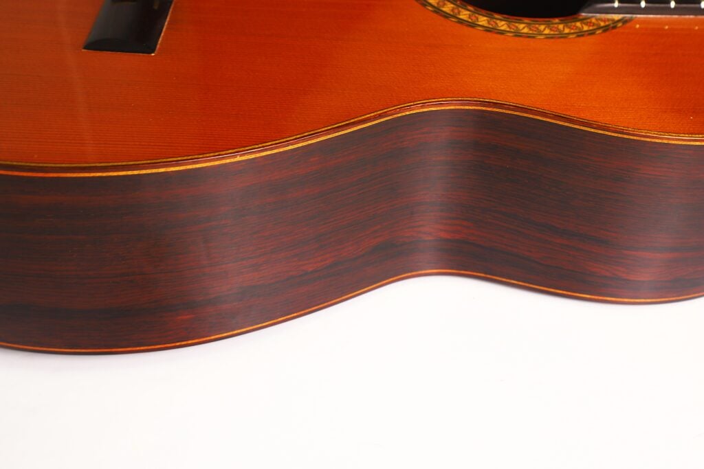 Nahaufnahme der Seite der 7-saitigen klassischen Gitarre „Melchor Rodriguez 1980 Ramirez Stil“, die die dunkle Holzmaserung, die glatte Oberfläche und einen Teil der Zierkante vor weißem Hintergrund hervorhebt. Nahaufnahme der Seite der 7-saitigen klassischen Gitarre „Melchor Rodriguez 1980 Ramirez Stil“, die die dunkle Holzmaserung, die glatte Oberfläche und einen Teil der Zierkante vor weißem Hintergrund hervorhebt.