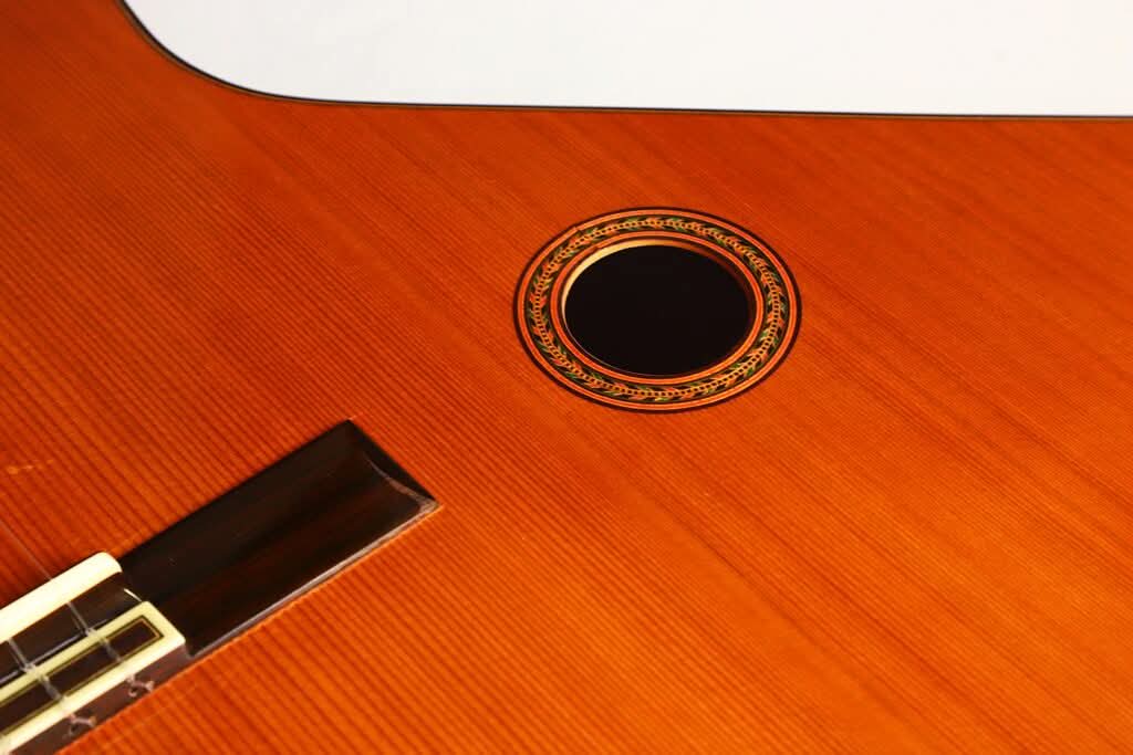 Nahaufnahme der „Gran Klassische Gitarre“ von Raimundo y Aparicio Corujo, bei der das Schallloch mit seiner dekorativen Rosette sowie ein Teil des Griffbretts vor dem Hintergrund einer polierten Holzoberfläche besonders hervorgehoben sind. Nahaufnahme der „Gran Klassische Gitarre“ von Raimundo y Aparicio Corujo, bei der das Schallloch mit seiner dekorativen Rosette sowie ein Teil des Griffbretts vor dem Hintergrund einer polierten Holzoberfläche besonders hervorgehoben sind.