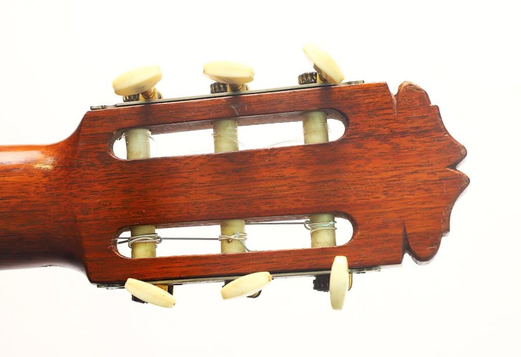 Nahaufnahme des Kopfes einer Flamenco-Gitarre von Manuel de la Chica aus dem Jahr 1954, die sechs cremefarbene Stimmwirbel und eine Holzoberfläche vor weißem Hintergrund zeigt. Nahaufnahme des Kopfes einer Flamenco-Gitarre von Manuel de la Chica aus dem Jahr 1954, die sechs cremefarbene Stimmwirbel und eine Holzoberfläche vor weißem Hintergrund zeigt.