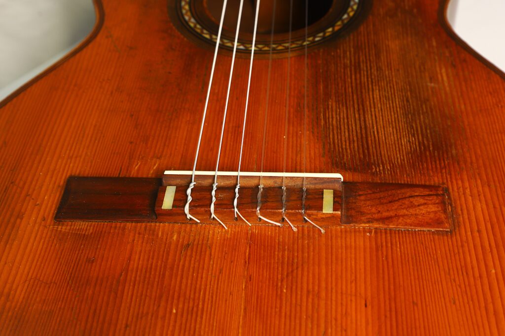 Nahaufnahme des Holzkörpers der „Ricardo Sanchis Nacher 1940 Augustin Barrios Klassische Gitarre“ mit Fokus auf Steg, Sattel und Saiten, einschließlich Detailansichten der Holzmaserung, der Saitenbefestigungen und der sichtbaren Abnutzungsspuren an der Oberfläche. Nahaufnahme des Holzkörpers der „Ricardo Sanchis Nacher 1940 Augustin Barrios Klassische Gitarre“ mit Fokus auf Steg, Sattel und Saiten, einschließlich Detailansichten der Holzmaserung, der Saitenbefestigungen und der sichtbaren Abnutzungsspuren an der Oberfläche.
