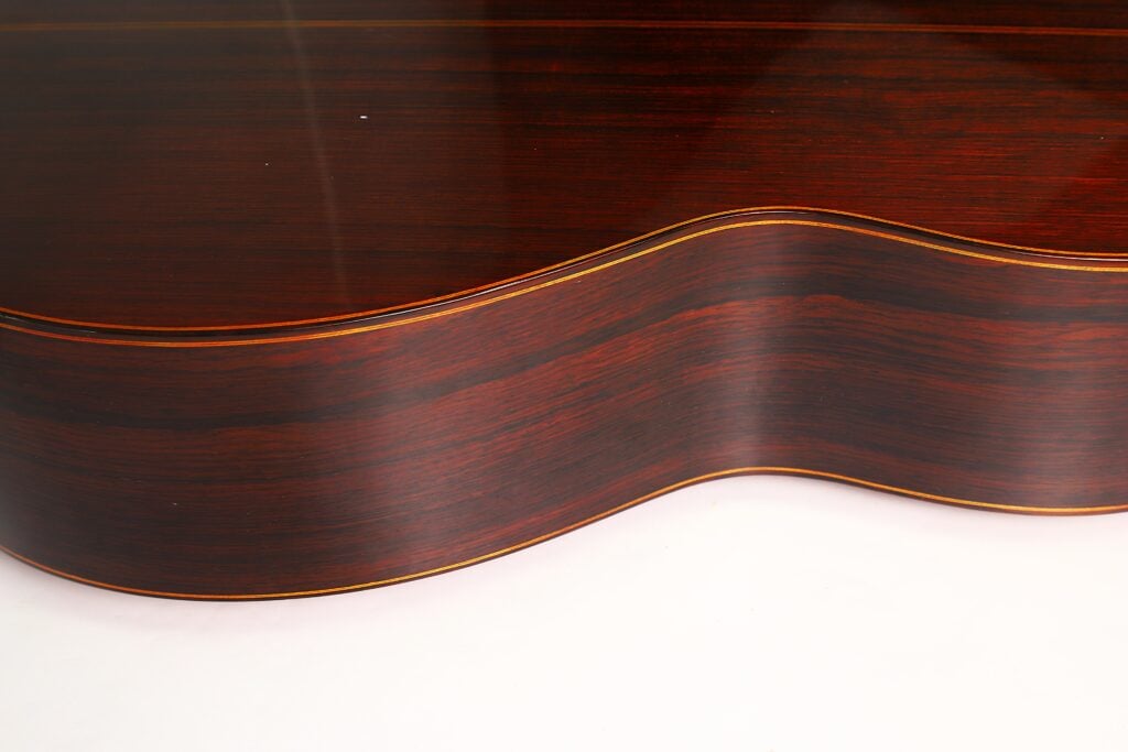 Nahaufnahme der Seite einer 7-saitigen klassischen Gitarre vom Typ „Ramirez Stil“ von Melchor Rodriguez aus dem Jahr 1980, die vor einem schlichten weißen Hintergrund die glänzende Lackierung, die dunkle Holzmaserung und die geschwungene Form hervorhebt. Nahaufnahme der Seite einer 7-saitigen klassischen Gitarre vom Typ „Ramirez Stil“ von Melchor Rodriguez aus dem Jahr 1980, die vor einem schlichten weißen Hintergrund die glänzende Lackierung, die dunkle Holzmaserung und die geschwungene Form hervorhebt.