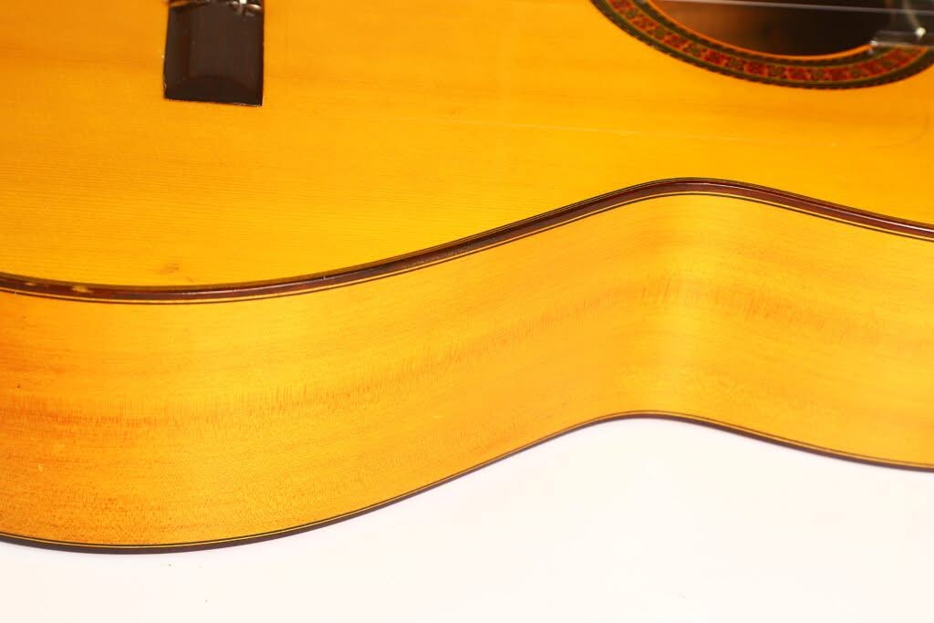 Nahaufnahme der Seite und des unteren Teils einer Flamenco-Gitarre von Sobrinos de Santos Hernandez aus dem Jahr 1974, die die Maserung des Holzes, die sanften Rundungen und die in der rechten oberen Ecke sichtbare Schalllochrosette hervorhebt. Nahaufnahme der Seite und des unteren Teils einer Flamenco-Gitarre von Sobrinos de Santos Hernandez aus dem Jahr 1974, die die Maserung des Holzes, die sanften Rundungen und die in der rechten oberen Ecke sichtbare Schalllochrosette hervorhebt.