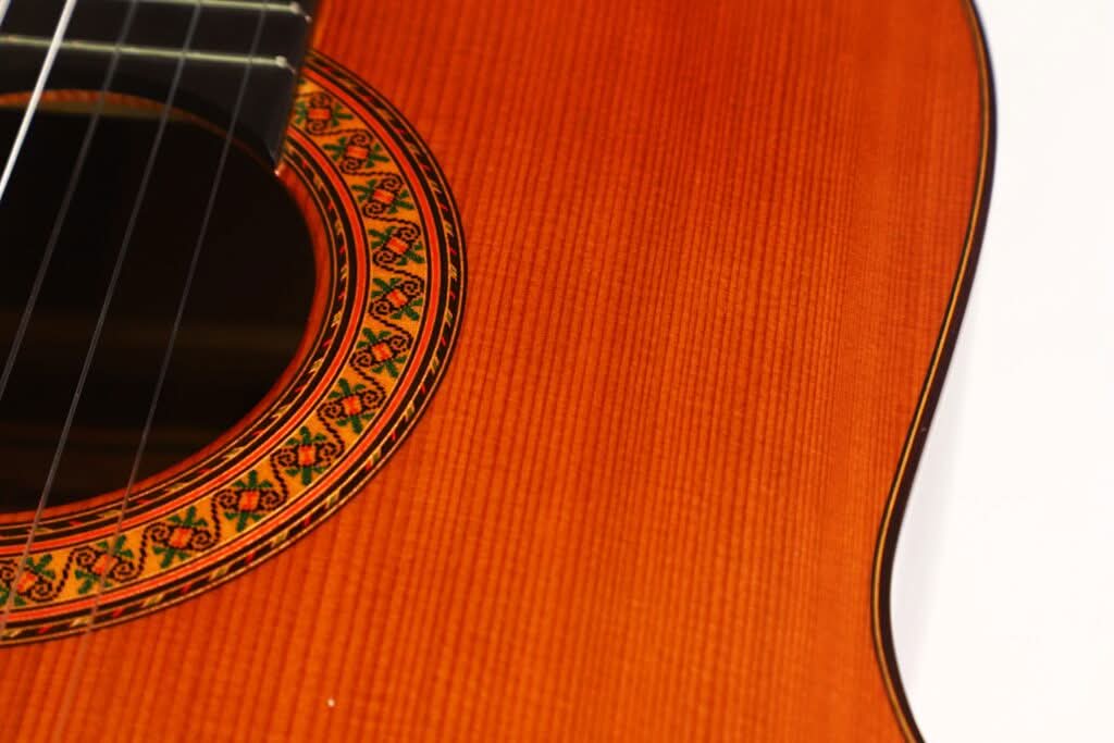 Nahaufnahme des Korpus der „Gran Klassische Gitarre“ von Raimundo y Aparicio Corujo, die die aufwendig gearbeitete Rosette, die das Schallloch umgibt, sowie die charakteristische Maserung der Gitarrendecke vor weißem Hintergrund hervorhebt. Nahaufnahme des Korpus der „Gran Klassische Gitarre“ von Raimundo y Aparicio Corujo, die die aufwendig gearbeitete Rosette, die das Schallloch umgibt, sowie die charakteristische Maserung der Gitarrendecke vor weißem Hintergrund hervorhebt.