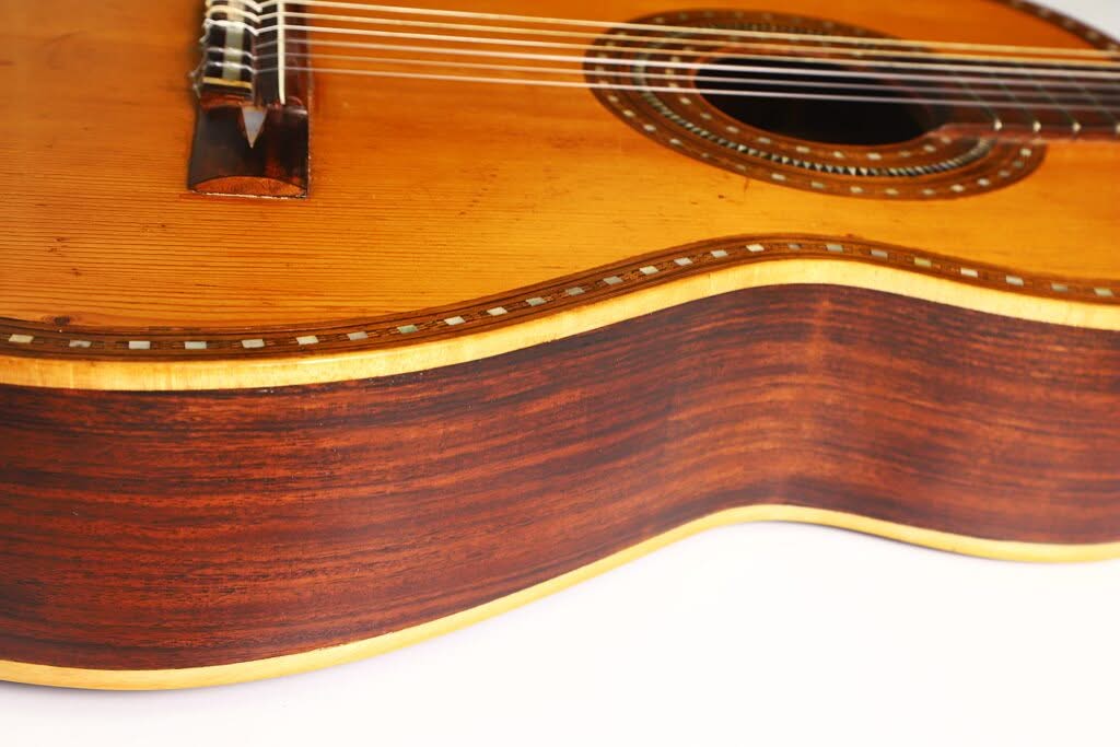 Nahaufnahme des Korpus der „Salvador Ibanez 1900 Klassische Gitarre“, die die Holzmaserung, die Saiten und die detailreiche Einlage am Schallloch zeigt, wobei der Fokus auf der Seite und der Decke vor einem weißen Hintergrund liegt. Nahaufnahme des Korpus der „Salvador Ibanez 1900 Klassische Gitarre“, die die Holzmaserung, die Saiten und die detailreiche Einlage am Schallloch zeigt, wobei der Fokus auf der Seite und der Decke vor einem weißen Hintergrund liegt.