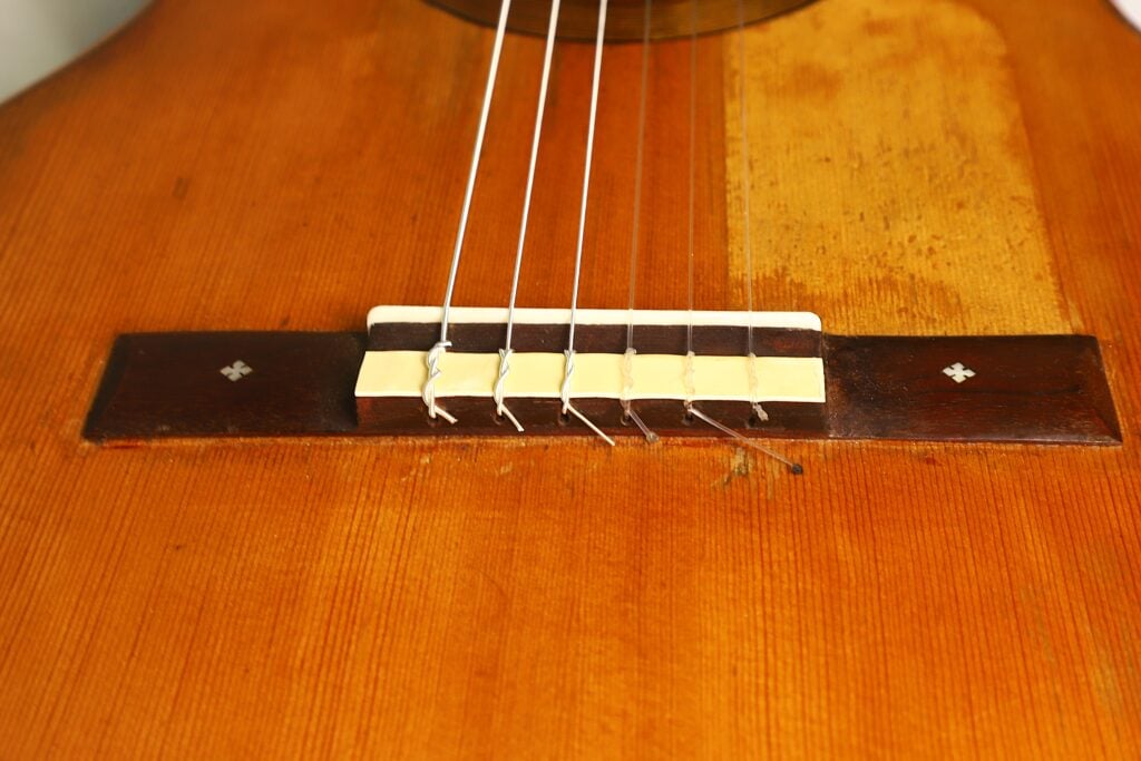 Nahaufnahme des Stegs der „José Ramirez 1932 Klassische Gitarre“, auf der sechs Saiten zu sehen sind, die an einem dunklen Holzsteg mit dekorativen Einlagen auf einem hellbraunen Holzkorpus befestigt sind. Nahaufnahme des Stegs der „José Ramirez 1932 Klassische Gitarre“, auf der sechs Saiten zu sehen sind, die an einem dunklen Holzsteg mit dekorativen Einlagen auf einem hellbraunen Holzkorpus befestigt sind.