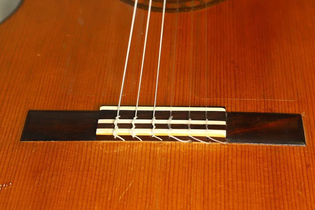Nahaufnahme des Stegs und der Nylonsaiten einer Flamenco-Gitarre von Manuel de la Chica aus dem Jahr 1954, bei der der Holzkorpus, der Steg und der Sattel besonders hervorgehoben werden. Nahaufnahme des Stegs und der Nylonsaiten einer Flamenco-Gitarre von Manuel de la Chica aus dem Jahr 1954, bei der der Holzkorpus, der Steg und der Sattel besonders hervorgehoben werden.
