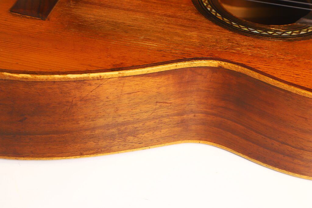Nahaufnahme der Holzseite und der Oberkante der „Ricardo Sanchis Nacher 1940 Augustin Barrios Klassische Gitarre“, die die satte braune Holzmaserung und die geschwungene Form vor weißem Hintergrund hervorhebt. Nahaufnahme der Holzseite und der Oberkante der „Ricardo Sanchis Nacher 1940 Augustin Barrios Klassische Gitarre“, die die satte braune Holzmaserung und die geschwungene Form vor weißem Hintergrund hervorhebt.