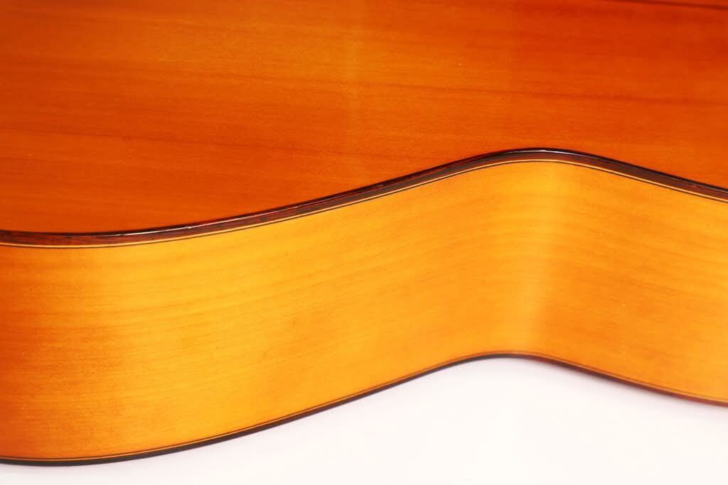 Nahaufnahme der Seite einer Jose Ramirez 1a Flamenco-Gitarre aus dem Jahr 1968, die sich durch ihren glatten, geschwungenen Holzkorpus mit einer naturbelassenen, hellbraunen Lackierung und dunklen Zierleisten auszeichnet. Nahaufnahme der Seite einer Jose Ramirez 1a Flamenco-Gitarre aus dem Jahr 1968, die sich durch ihren glatten, geschwungenen Holzkorpus mit einer naturbelassenen, hellbraunen Lackierung und dunklen Zierleisten auszeichnet.