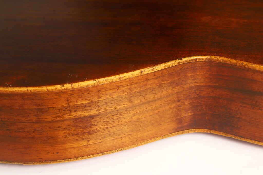 Nahaufnahme der Seite einer „Ricardo Sanchis Nacher 1940 Augustin Barrios Klassische Gitarre“, die die Holzmaserung, die abgenutzten Kanten und die Lackierung zeigt. Die Beleuchtung hebt die Rundungen und die Textur dieses klassischen Instruments hervor. Nahaufnahme der Seite einer „Ricardo Sanchis Nacher 1940 Augustin Barrios Klassische Gitarre“, die die Holzmaserung, die abgenutzten Kanten und die Lackierung zeigt. Die Beleuchtung hebt die Rundungen und die Textur dieses klassischen Instruments hervor.