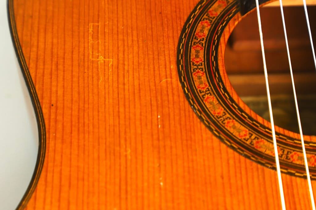 Nahaufnahme der Flamenco-Gitarre „Manuel de la Chica 1954“ mit Fokus auf die Holzmaserung, die dekorative Rosette um das Schallloch und die weißen Saiten. Nahaufnahme der Flamenco-Gitarre „Manuel de la Chica 1954“ mit Fokus auf die Holzmaserung, die dekorative Rosette um das Schallloch und die weißen Saiten.