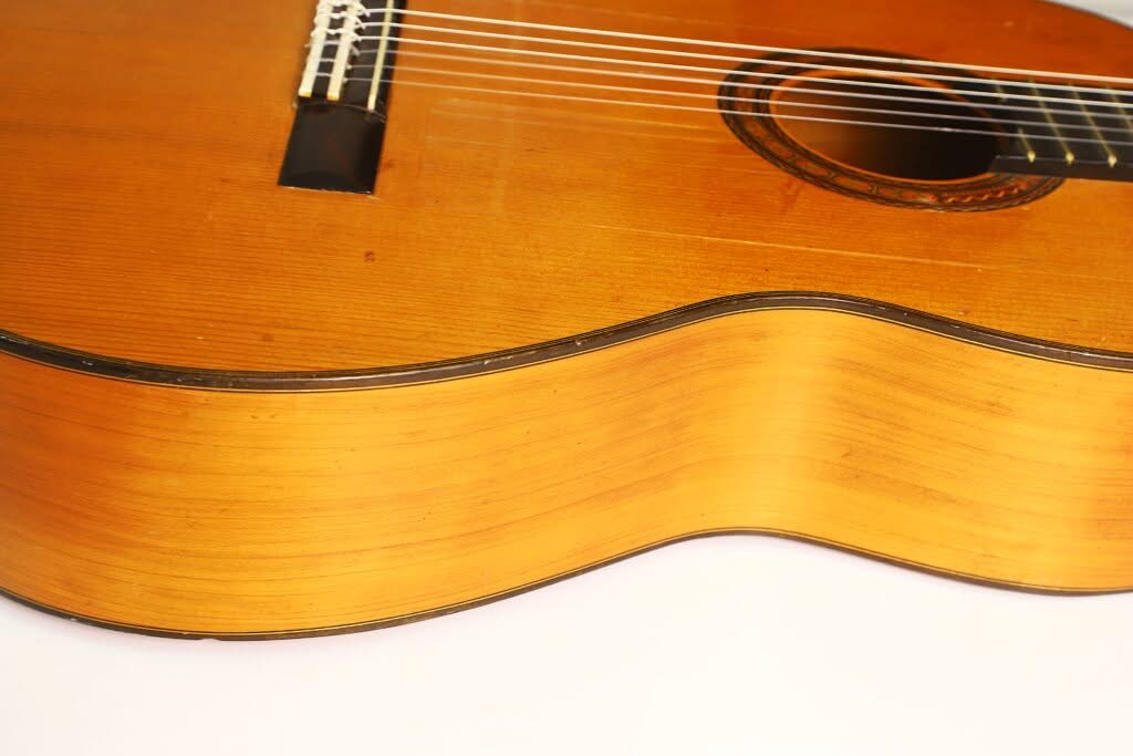 Nahaufnahme des Korpus einer Flamenco-Gitarre von Manuel de la Chica aus dem Jahr 1954, die die Holzmaserung, die Saiten und das Schallloch mit ihrer warmen braunen Lackierung vor weißem Hintergrund hervorhebt. Nahaufnahme des Korpus einer Flamenco-Gitarre von Manuel de la Chica aus dem Jahr 1954, die die Holzmaserung, die Saiten und das Schallloch mit ihrer warmen braunen Lackierung vor weißem Hintergrund hervorhebt.