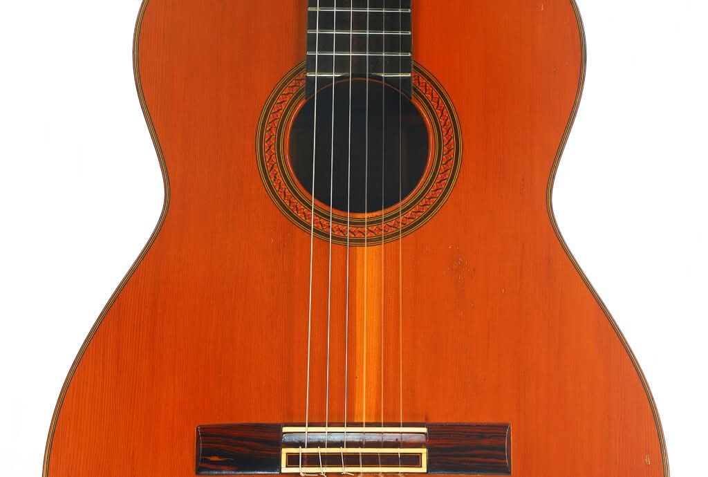 Nahaufnahme des vorderen Korpus und der Saiten der „Klassische Gitarre“ von Antonio Duran aus dem Jahr 1974, mit Holzoberfläche, Schallloch und aufwendig gestaltetem Rosettenmuster vor weißem Hintergrund. Nahaufnahme des vorderen Korpus und der Saiten der „Klassische Gitarre“ von Antonio Duran aus dem Jahr 1974, mit Holzoberfläche, Schallloch und aufwendig gestaltetem Rosettenmuster vor weißem Hintergrund.