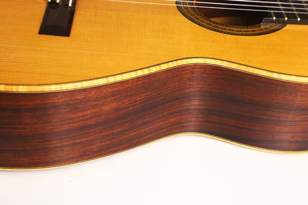 Nahaufnahme der Seite und des Korpus der „Hauser Klassische Gitarre“ von Alastair McNeil aus dem Jahr 1997, die die Maserung des Holzes und die Zierleisten hervorhebt; oben sind ein Teil der Saiten und das Schallloch zu sehen. Nahaufnahme der Seite und des Korpus der „Hauser Klassische Gitarre“ von Alastair McNeil aus dem Jahr 1997, die die Maserung des Holzes und die Zierleisten hervorhebt; oben sind ein Teil der Saiten und das Schallloch zu sehen.