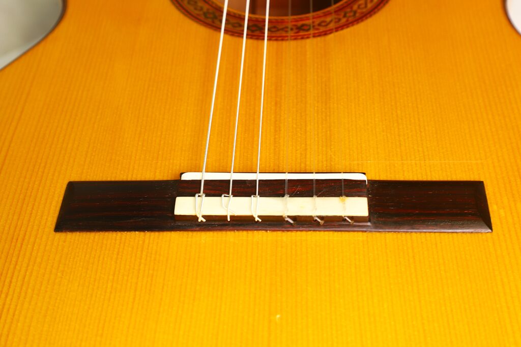 Nahaufnahme des Stegs und der Saiten der Jose de Jesus 2024 Blanca Modell Reyes Flamenco-Gitarre mit einem Holzkorpus in einem warmen Gelbton. Nahaufnahme des Stegs und der Saiten der Jose de Jesus 2024 Blanca Modell Reyes Flamenco-Gitarre mit einem Holzkorpus in einem warmen Gelbton.