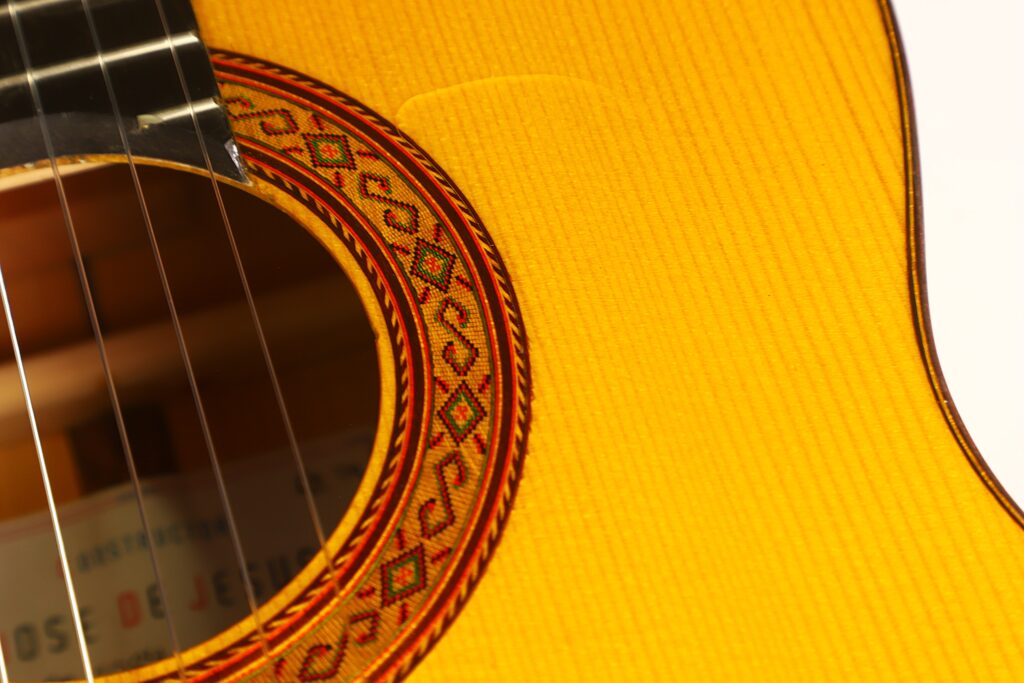 Nahaufnahme des Schalllochs und des filigranen Rosettenmusters auf dem Holzkorpus der Jose de Jesus 2024 Blanca Modell Reyes Flamenco-Gitarre, wobei links teilweise die Saiten zu sehen sind. Nahaufnahme des Schalllochs und des filigranen Rosettenmusters auf dem Holzkorpus der Jose de Jesus 2024 Blanca Modell Reyes Flamenco-Gitarre, wobei links teilweise die Saiten zu sehen sind.