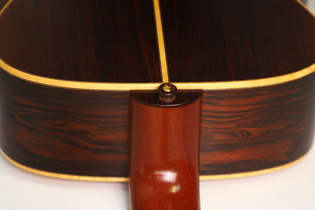 Nahaufnahme des Rückens der Lakewood M-54 Akustikgitarre aus dem Jahr 1998, mit einem glatten Halsansatz aus Holz, dunkel poliertem Holz mit heller Einfassung an den Kanten und einem goldenen Gurtknopf am unteren Rand. Nahaufnahme des Rückens der Lakewood M-54 Akustikgitarre aus dem Jahr 1998, mit einem glatten Halsansatz aus Holz, dunkel poliertem Holz mit heller Einfassung an den Kanten und einem goldenen Gurtknopf am unteren Rand.